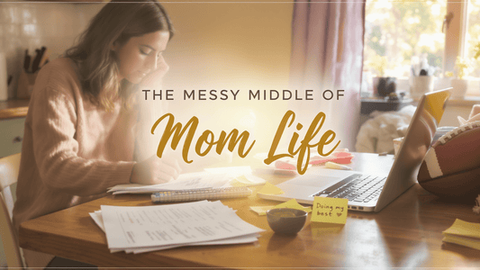 A woman writing at a cluttered table, embodying the messy middle of motherhood with notes and a laptop.