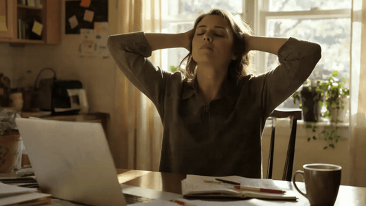 Hustle to Burnout: A mom sitting at her desk, surrounded by papers, laptop open, elbows up, hands resting on her head